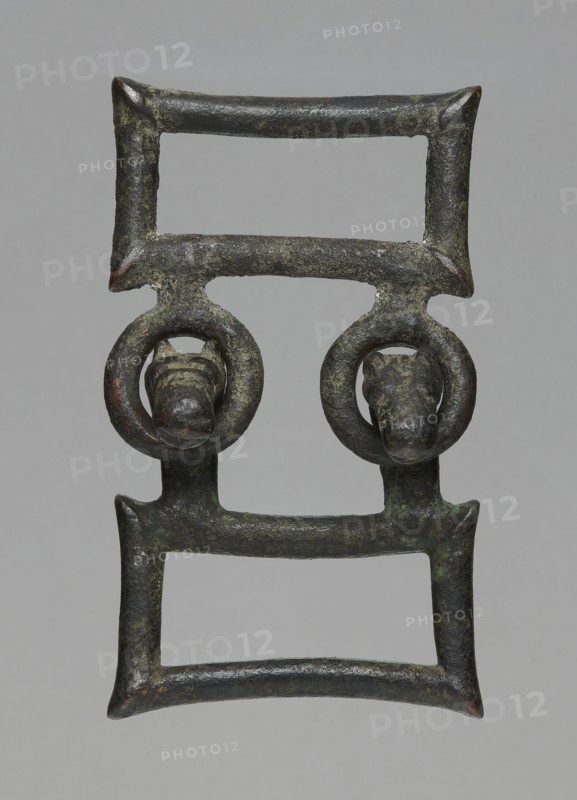 Buckle, bronze; two-part rectangular form with joints; one part with hooks shaped as zoomorphic animal heads; decoration; 7th century BC, Etruria; length 13 cm total, height 7.5 cm, thickness 4 cm, weight 0.374 kg, length 7.2 cm single element., An aged metal object with a rectangular, ladder-like form composed of two parallel bars at top and bottom connected by vertical segments, featuring a pair of circular loops suspended from the middle bar each encircling a small sculpted animal head; the surface shows a weathered, rough texture with patches of dark green and brown patina, along with areas of darker gray and hints of reddish-brown.