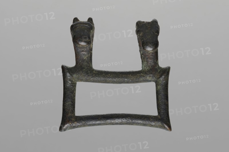 Buckle, Bronze buckle consisting of two parts in a shape similar to rectangles with joints. One part of the buckle has hooks in the form of zoomorphic decoration animal heads. 7th century BC, Etruria. Decoration. Length 13 cm total, height 7.5 cm, thickness 4 cm, weight 0.374 kg, length 7.2 cm single element., An aged metal object with a rectangular open center and a slightly curved lower bar, showing a rough, pitted surface in dark green, dark brown, black, and hints of lighter green patina, topped by a pair of short vertical posts each ending in a rounded animal-like head with small ears and snouts, all set against a plain light gray background.