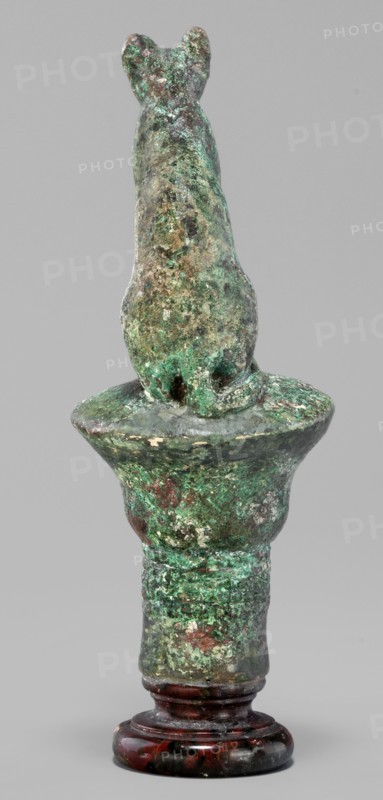 Cat sitting on a papyrus column, cats; Scen.Gal.Eg.; capitals; Egyptian art; 664–332 BC Late Period, Egypt; figurine; height 11 cm, width 4.5 cm, weight 0.314 kg., An upright sculptural object resembling an animal figure perched on a rounded pedestal, shown from behind against a plain gray background, with an overall weathered surface showing a mottled mix of green, brown, and turquoise patina, darker brown and reddish areas, and some lighter worn spots; the base appears glossy dark reddish-brown with black veining.