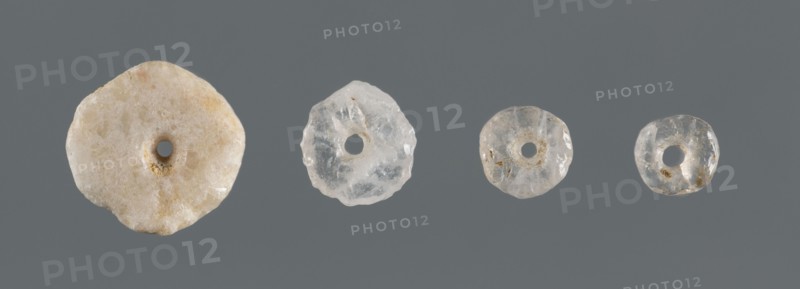 Set of four disc-shaped beads of different sizes, unknown northern Mesopotamian workshop; beads, disc-shaped, jewellery; likely from children's graves; ca. 2600–ca. 2350 BC, Early Jazira III period Early Dynastic III; Northern Mesopotamia Khabur basin, Tell Rad Shakra, Northeastern Syria; decoration., A row of four round, donut-shaped translucent objects arranged against a solid grey background, each with a central hole and irregular, slightly rough edges; the leftmost piece is larger and milky off-white with beige and pale tan speckles and a crystalline texture, the next is a clear to cloudy pale white with subtle internal fractures, the next is slightly smaller and translucent with faint beige inclusions and a glossy surface, and the rightmost is the smallest, mostly clear with small tan flecks and a shiny, irregular surface.