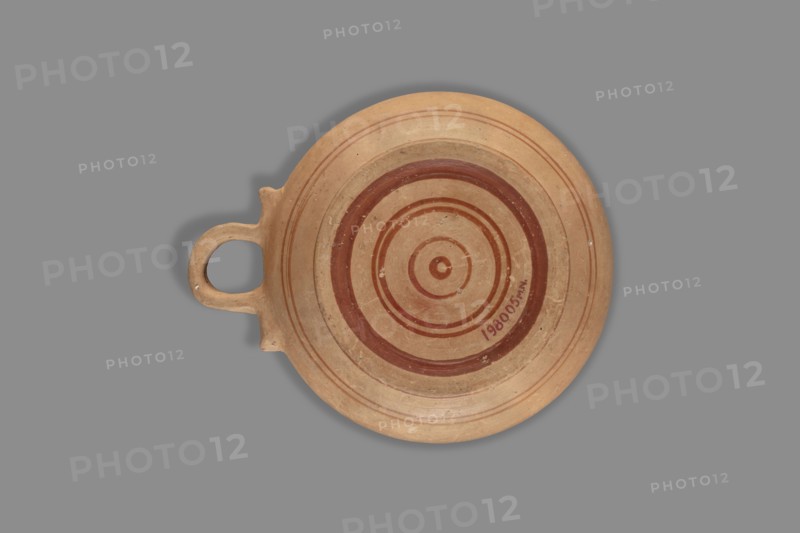 Exaleiptron kothon on a low, conical foot. Ribbon-shaped base; upper part and inner surface covered with red varnish thin, uneven layer; outer part of foot covered with red varnish. Band decoration painted on clay with delicate red/orange lines; decoration includes concentric circles on the bottom and a main ornament of smaller, narrower leaves. Smooth surface with visible turning traces; fine-grained clay with visible pyroxene. Glued artifact with visible conservation work; large losses in varnish and impurities inside. Clay color: Red 10 R 5/4–10 R 5/8. Attributed to Corinth; dated to the 2nd half of the 6th century BC. Vessel kothon, exaleiptron. Dimensions: height 5.5 cm, width 17.4 cm, depth 14.5 cm; weight 0.304 kg., A round shallow clay dish with a single loop handle is shown against a solid gray background; the dish is light beige with concentric painted rings in reddish-brown and darker brown tones, and the surface shows small chips and wear spots with slight variations in bei...