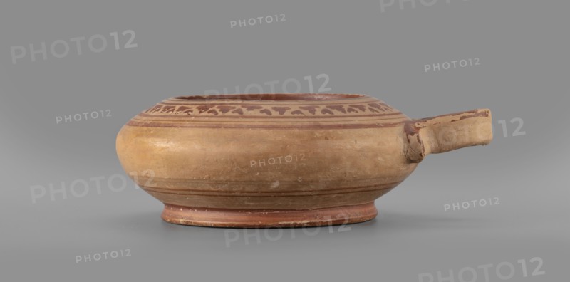 Exaleiptron kothon on a low, conical foot; ribbon-shaped base; upper part and inner surface covered with red varnish thin, uneven layer; outer part of foot covered with red varnish. Band decoration painted on clay with delicate orange/red lines and concentric circles on the bottom. Main decorative ornament consists of smaller, narrower leaves; plant and geometric motifs. Smooth surface with visible traces of turning; fine-grained clay with visible pyroxene. Glued artifact with visible conservation work; large losses in varnish and impurities inside. Clay color: 10 R 5/4 weak red, 10 R 5/8 red. Attributed to Corinth; 2nd half of the 6th century BC. Vessel kothon, exaleiptron. Height 5.5 cm, width 17.4 cm, depth 14.5 cm, weight 0.304 kg., A shallow, rounded ceramic bowl with a short spout on one side resting on a low circular foot; the bowl is primarily tan and light brown with darker reddish-brown painted bands and a decorative band of small abstract shapes near the rim, the spout showi...
