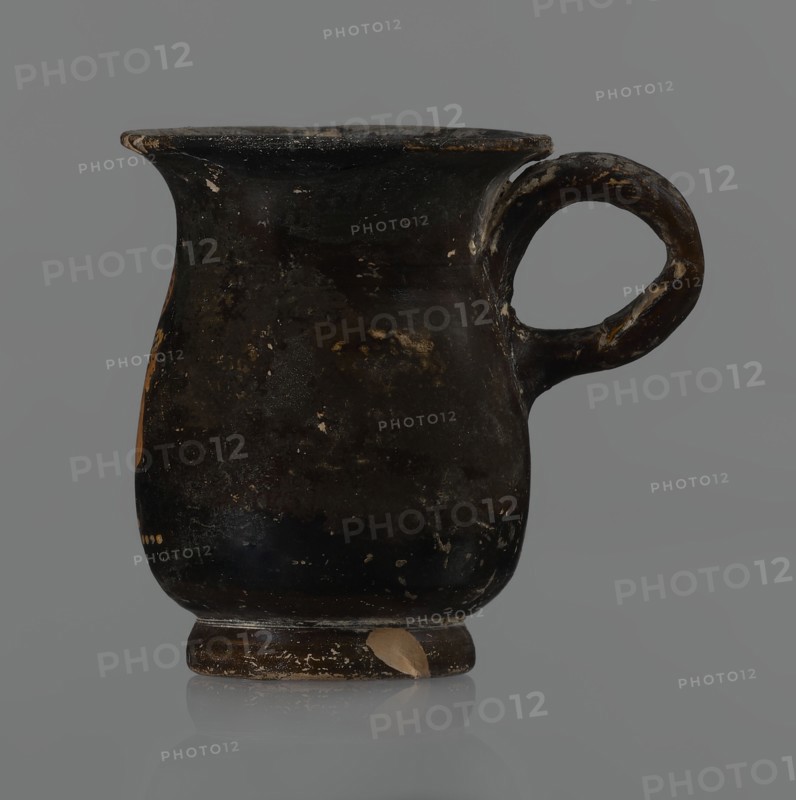 Jug, red-figure pottery, Greek pottery, mermaid mythological, Athens Greece. A small jug with a low foot recessed at the bottom. The edges of the foot are slightly rounded and tapered upwards. The body is slightly wider than the foot, rounded at the bottom, almost cylindrical in the lower half, and gently tapering in the upper half, merging into a widely flared outward spout. Below the spout is a vertically mounted, cylindrical, slightly flattened handle, bent into a circle. Light orange clay, black shiny firnis. On the body, the mermaid is in right profile, with wings raised upwards the left wing at the back, protruding above the right. On the mermaid's head is a diadem marked with white dots. Feathers marked with lines—long and thin at the wingtips and tail; thin and thick elsewhere. The image is emphasized from the bottom with a row of dots. The side of the foot is chipped, the rim edge is slightly chipped, and the firnis is abrasion-stripped. Part of the rim is filled with plaster....