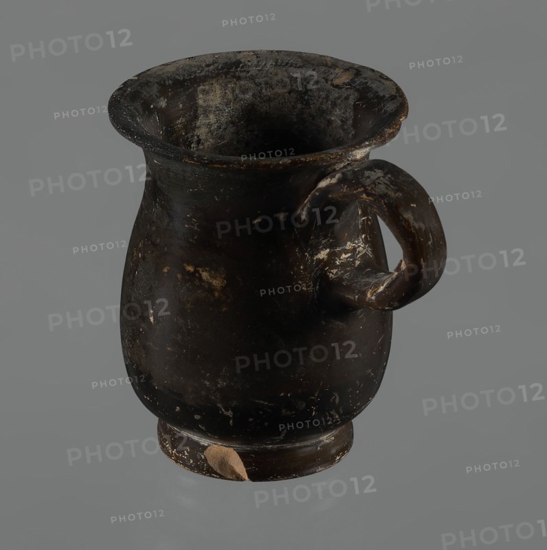 Jug, red-figure pottery, Greek pottery, mermaid mythological, Athens Greece. A small jug with a low foot recessed at the bottom. The edges of the foot are slightly rounded and tapered upwards. The body is slightly wider than the foot, rounded at the bottom, almost cylindrical in the lower half, and gently tapering in the upper half, merging into a widely flared outward spout. Below the spout is a vertically mounted, cylindrical, slightly flattened handle, bent into a circle. Light orange clay, black, shiny firnis. On the body, the mermaid is in right profile, with wings raised upwards the left wing at the back, protruding above the right. On the mermaid's head is a diadem marked with white dots. Feathers marked with lines—long and thin at the wingtips and tail; thin and thick elsewhere. The image is emphasized from the bottom with a row of dots. The side of the foot is chipped, the rim edge is slightly chipped, and the firnis is abrasion-stripped. Part of the rim is filled with plaster...