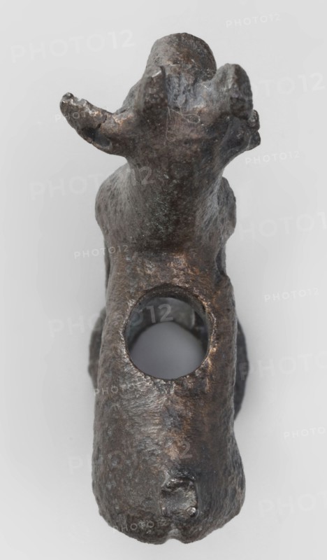 Emblem in the form of a figurine of a standing goat figurine of a standing gazelle with slightly bent legs on a ring-shaped base. Through the center of the animal's body runs a sleeve-shaped hole aligned with the hole in the ring, allowing the figurine to be mounted on, for example, a wooden handle. The animal's protruding ears contain holes that could have been used for hanging ornaments, e.g., bells. Luristan bronze, unknown Luristan workshop, 1st half of the 1st millennium BC Iron Period II, Luristan, Iran. Materials: bronze; dimensions: height 6.5 cm, width 4.8 cm, depth 1.7 cm., A dark brown and black metal object shaped like an animal form is shown from above against a light gray background, with a rounded head and two upward curving ear or horn-like protrusions, a textured surface with irregular dents and scratches, a large circular hole through the middle of its body, and a few areas of lighter brownish highlights and subtle greenish patina.