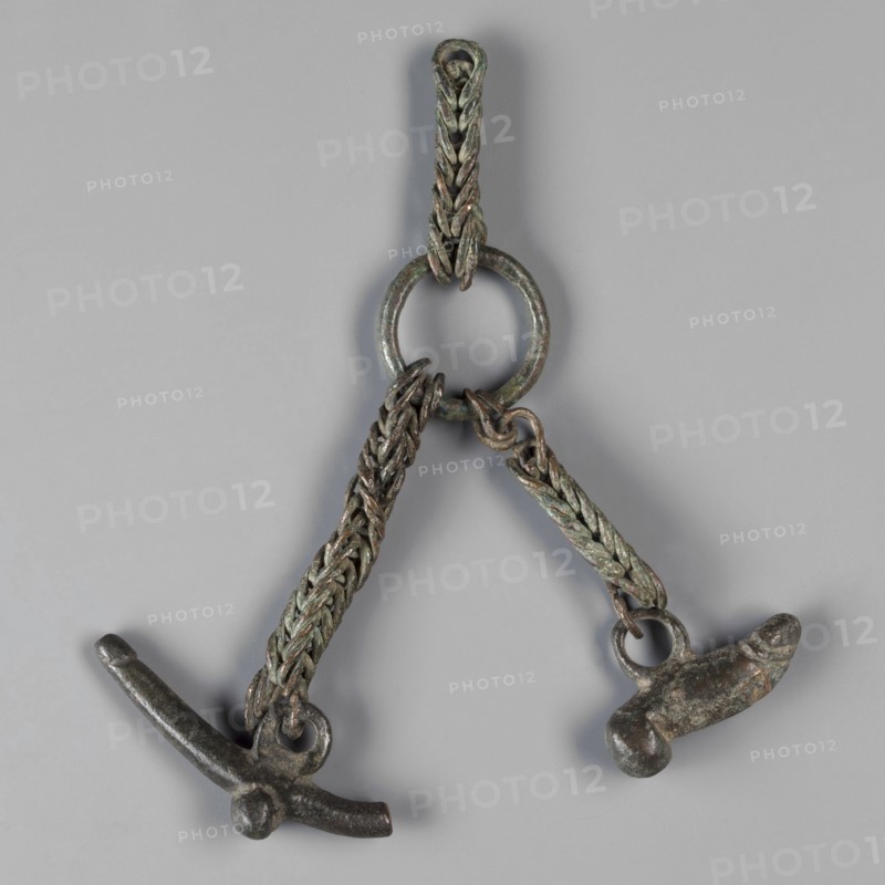 Pendant with phalluses, phalluses, pendants, amulets - body parts, 1st century, Italy, amulet, weight 50 g, An object consisting of a circular ring with two braided chains hanging from it, each chain ending in a small metal pendant shaped with rounded and elongated forms; the metal shows a patina with greenish and brownish tones and darker gray areas, and the background is a uniform light gray.