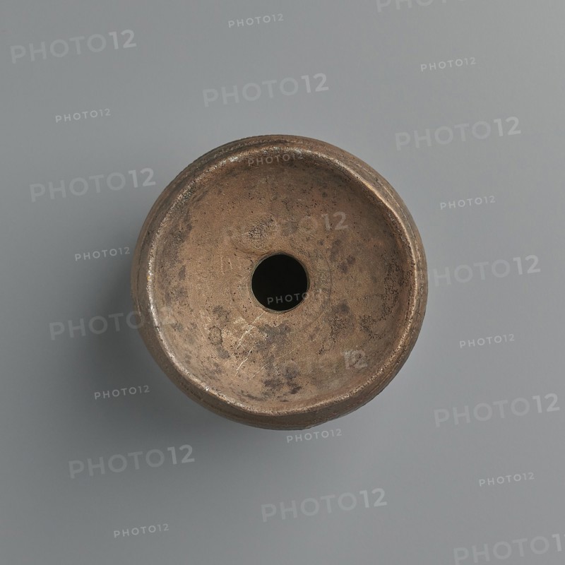 Aryballos, 2nd–1st century BC, Sicily Segesta? incorrect information?, Etruria possible origin, vessel aryballos, weight 0.11 kg, height 5.4 cm, diameter 5 cm, A round, bowl-shaped object viewed from above with a central circular hole, showing a textured, worn surface in shades of brown and tan with darker brown spots and patches, resting on a smooth background in shades of gray and bluish-gray, with subtle shadowing beneath the object.