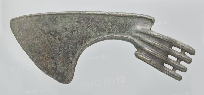 Battle axe, unknown Luristan workshop. Battle axe with a low cylindrical socket with a round hole, from which grows a blade terminating in a semicircular cutting edge set at ~45° to the axis of the shaft. The base of the blade protrudes above the socket; its upper edge is sloping and straight with a slight bulge in the middle, while the lower edge is shorter and slightly obtuse arc-shaped. Four identical blunt-tipped spikes project from the butt, connected vertically at mid-length; the spikes extend into side ribs on both sides of the socket — the upper two terminate at the blade base, the third extends into its lower edge, and the fourth runs around the lower edge of the socket, reinforcing it. Luristan, Iran; 2nd half of the 2nd millennium BC. Weapon. Weight 334 g; height 10.2 cm; length 19 cm; thickness 3.2 cm., An aged metal object with a broad, roughly triangular blade area showing a mottled surface in dark gray, brown, and patches of green, connected to a curved neck that leads t...