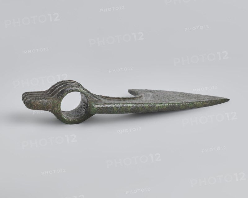 Battle axe, unknown Luristan workshop. Battle axe with a low cylindrical socket with a round hole, from which grows a blade terminating in a semicircular cutting edge set at ~45° to the shaft axis. The base of the blade protrudes above the socket; its upper edge is sloping and straight with a slight central bulge, the lower edge is shorter and a slightly obtuse arc. Four identical blunt-tipped spikes protrude from the butt, connected vertically at mid-length. The spikes extend into side ribs on both sides of the socket: the upper two terminate at the base of the blade, the third extends into its lower edge, and the fourth runs around the lower edge of the socket, further reinforcing it. Luristan bronzes; weapon. 2nd half of the 2nd millennium BC. Iran; Luristan. Weight 334 g; height 10.2 cm; length 19 cm; thickness 3.2 cm., A single metallic object lies on a pale gray background, showing a long flat blade that tapers to a pointed tip and a rounded section with a circular hole near the ...