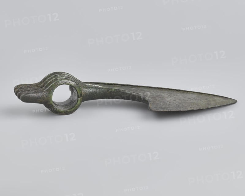 Battle axe, unknown Luristan workshop. Battle axe with a low cylindrical socket with a round hole, from which grows a blade terminating in a semicircular cutting edge set at about 45° to the shaft axis. The base of the blade protrudes above the socket; its upper edge is sloping and straight with a slight central bulge, the lower edge is shorter and slightly obtuse-arched. Four identical blunt-tipped spikes protrude from the butt, connected vertically at mid-length; the spikes extend into side ribs on both sides of the socket — the upper two terminate at the base of the blade, the third extends into its lower edge, and the fourth runs around the lower edge of the socket for reinforcement. Luristan, Iran; 2nd half of the 2nd millennium BC. Bronze weapon. Weight 334 g; height 10.2 cm; length 19 cm; thickness 3.2 cm., A metal object with a long flat blade-like section tapering to a pointed end lies horizontally on a pale gray background; the opposite end forms a rounded ring with a hollow ...
