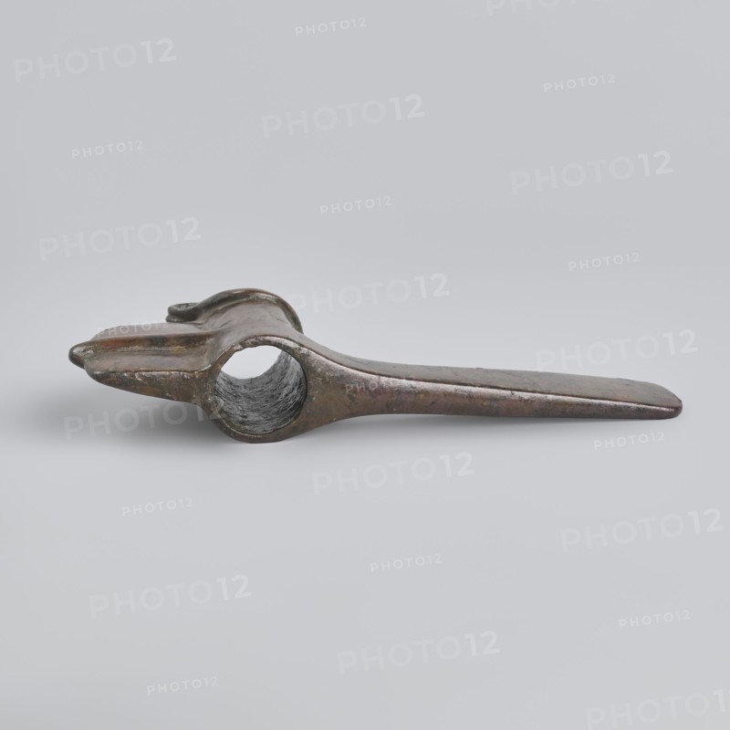 Battle axe, Eastern Gallic Scenography, 2nd half of 3rd millennium BC, Western Iran, weapon, weight 280 g, A single elongated metallic object rests on a light gray background, showing a worn, slightly curved form with a hollow circular opening near one end and a flattened tapered end at the other; the surface displays a mottled mix of dark bronze, brown, and hints of green patina with areas of darker gray inside the hollow and subtle shiny reflections on the outer surfaces.