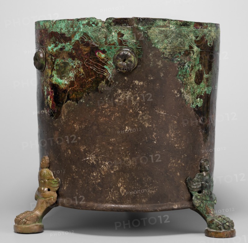Cista container decorated with a depiction of the Dioscuri and Athena, Cylindrical vessel made of sheet metal, decorated with an engraved depiction of the Dioscuri and Athena, 8 figures preserved. Under the rim and at the lower edge are bands of engraved plant decoration. Concave bottom. Legs in the shape of predator's paws, ending in sirens. Slightly convex lid, made of sheet metal, decorated with an engraved depiction of two hippocampi. Trace of a handle in the centre. 4th century BC, Praeneste Italy. Vessel; metal; Cista on 3 legs. Weight 1.132 kg; height 24.5 cm; diameter 20 cm bottom, 22 cm top., A cylindrical metal vessel with a worn, mottled surface showing dark brown and green patina, with areas of bright green corrosion and traces of reddish and black discoloration near the rim; the rim has a rough edge and several raised round rivet-like features. The lower edge rests on a group of three sculpted feet that appear as stylized animal or creature forms with a mix of brown, green...
