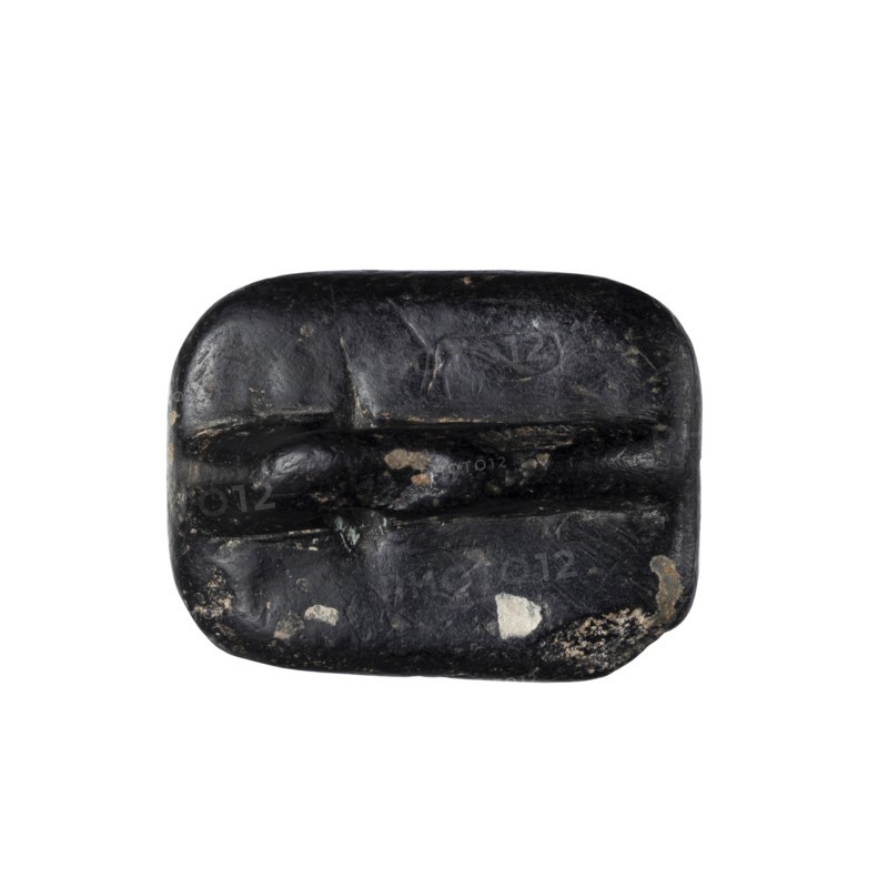 Rectangular stamp seal with a handle in the form of a protruding ridge along the axis of the seal with a crosswise drilled hole. On a rectangular base with rounded corners is a schematic representation of a demon, in human form and with wings instead of arms. Unspecified filler elements are on the sides of the demon. c. 1650–c. 1350 BC; Late Bronze Period. Northwest Syria place of manufacture, Near East place of find. Material: seal. Dimensions: height 1.7 cm, width 4.2 cm, depth 3.2 cm., A dark rounded rectangular object with a smooth, slightly worn surface showing a few rough patches and chips in lighter tones; the object is primarily black with small areas of gray, off-white, and beige where the surface is worn away, and it has shallow carved or indented grooves across its face giving it a segmented appearance.