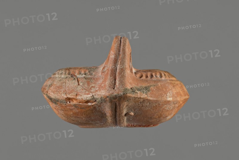 Oil lamp, 5th century, Carthage, oil lamp, height 5.3 cm, width 8.7 cm, depth 13.6 cm, A small, worn terracotta object in shades of reddish-brown and orange sits against a gray background; it has a rounded, boat-like body with a central raised vertical ridge forming a split peak, and a textured rim with a pattern of small indentations. The surface shows areas of lighter beige and pale gray weathering and deposits, with visible cracks and rough patches, and the underside displays smoother, slightly darker brown tones. Overall the piece appears aged with uneven coloration including reddish-brown, orange, beige, pale gray, and darker brown.