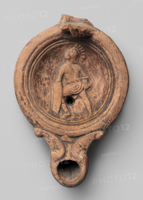 Lamp decorated with a depiction of a gladiator so-called Samnite, unknown Roman workshop, Scen.Gal.Class., gladiators iconography, lighting, oil lamps, single-burner lamps, 2nd half of the 2nd century, Roman Empire 27 BC–476, oil lamp, weight 0.078 kg, An oval terracotta object in shades of reddish-brown and dark brown with weathered surface texture shows a recessed circular field containing a low-relief figure wearing a draped garment and a helmet-like headpiece, standing with one leg forward and holding a rectangular shield and a short object; concentric raised rings surround the recessed field, and there is a central round hole within the scene. The rim and body exhibit worn areas and darker patina, with a small protrusion at the top and a narrow elongated spout at the bottom that forms a rounded opening; the lower handle area features molded scroll-like shapes and similar worn coloration.
