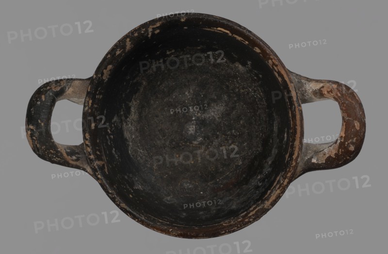 Miniature skyphos. Reddish clay, black-brown firnis poorly covering the outside on the foot, at the bottom of the body, the outer parts of the handles, the rim and the inside. The zone between the handles is white-ground, black-figure. Decoration: A - seated human figure in right profile; B - standing human figure facing right and quadruped facing left. 1st quarter of 5th century BC, Attica. Vessel, skyphos; height 4 cm, width 5.2 cm., unknown Attic workshop. Materials/technique: white-ground and black-figure pottery; miniature vessel; palmette ornament., A top-down view of a round, shallow bowl or pot with two opposing handles, showing a worn, rough interior and exterior surface; the object displays a mix of black, dark gray, brown, beige, and hints of off-white, with patches where the darker surface has chipped to reveal lighter underlying material, set against a plain gray background.