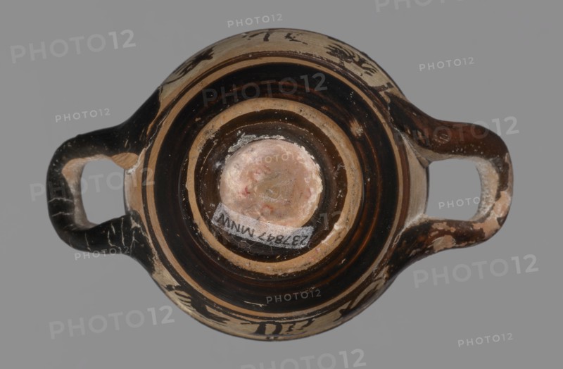 Miniature skyphos. Reddish clay, black-brown firnis poorly covering the outside on the foot, at the bottom of the body, the outer parts of the handles, the rim and the inside. The zone between the handles is white-ground, black-figure. Decoration: A - seated human figure in right profile; B - standing human figure facing right and quadruped facing left., Attic workshop; 1st quarter of 5th century BC, Attica Athens, Ancient Greece. Vessel skyphos, height 4 cm, width 5.2 cm. Palmettes ornament; miniature vessel; white-ground and black-figure pottery., A top-down view of a small two-handled vessel with a rounded body and a central circular base area, showing concentric bands of dark brown and beige on the interior and exterior surfaces; the handles extend horizontally on either side and show worn areas revealing lighter beige beneath a dark brown surface; the central base has a pale, slightly pinkish beige patch with some irregular discoloration and residue; the vessel surface shows chipp...