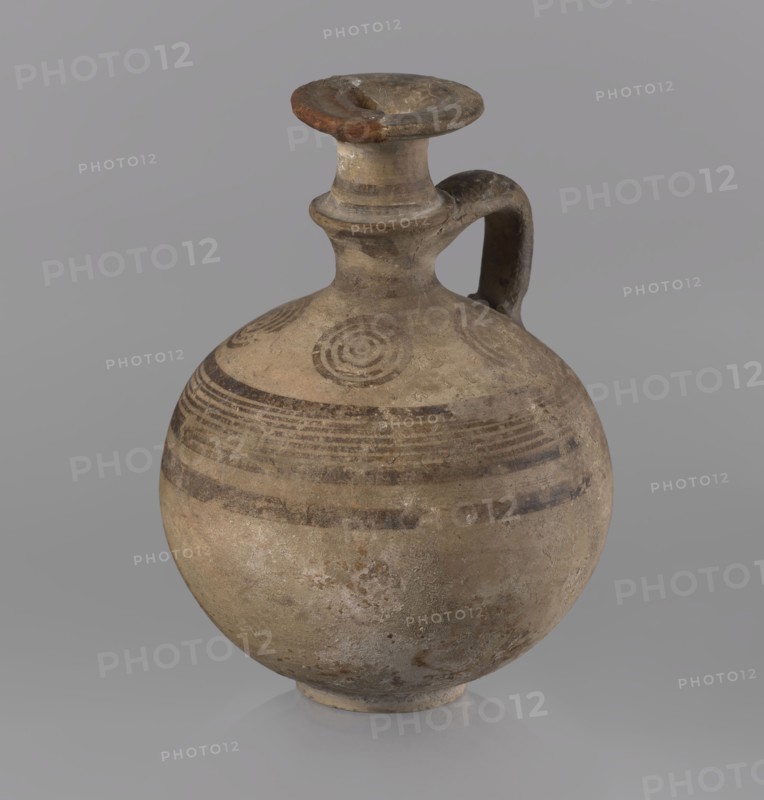 Jug, Cypriot, painted pottery, 850–750 BC Cypriot Geometric Period III. Jug with a flat foot, spherical body, high narrow neck with a strongly tilted spout, and a small vertical handle supported by arms and a ring on the neck. Decoration: above the maximum diameter a black band and a set of five parallel lines framed by thicker bands; the arms bear four sets of concentric circles arranged symmetrically on the sides of the vessel's axis; handle decorated with transverse lines. Medium: painted pottery. Function: vessel jug. Dimensions: height 10.5 cm, width 7.8 cm., A rounded ceramic vessel with a narrow neck and a rounded lip, featuring a single curved handle attached from the neck to the shoulder, resting on a small circular base; the surface shows a weathered, rough texture with areas of wear and pitting, and painted concentric circular motifs and horizontal banding around the shoulder; visible colors include beige, tan, brown, gray, and hints of darker brown in the painted decoration...