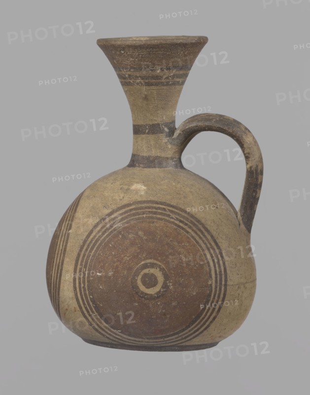 Cypriot jug, Cypriot pottery, Cypriot Bichrome Ware III. Jug on a low, straight foot. Globular body, flattened at the bottom. Quite narrow neck, funnel-shaped spout. Slightly flattened handle attached to the base of the neck and the upper part of the body. Painted decoration in stripes and linear patterns, in red-brown and black fading to brown. Near the spout is a wide red band, below it three black lines, the first adjacent to the red band. At the lower part of the neck, above the handle attachment, a narrow black band; another narrow black band at the base of the neck. The belly is decorated with three identical shooting target motifs on the front and sides: a wide red circular band with a black dot and a circle in the center, surrounded on the outside by four black-brown circles, the first adjacent to the red band. The handle is unpainted on the vessel side, black on the top, with light horizontal lines. The foot on the side is black. On the bottom, two concentric circles with a ce...