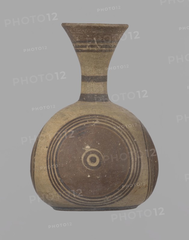 Cypriot jug, Jug on a low, straight foot. Globular body, flattened at the bottom. Quite narrow neck, funnel-shaped spout. Slightly flattened handle attached to the base of the neck and the upper part of the body. Painted decoration in stripes and linear patterns, in red-brown and black fading to brown. Near the spout is a wide red band, below it three black lines, the first adjacent to the red band. At the lower part of the neck, above the handle attachment, a narrow black band; another narrow black band at the base of the neck. The belly is decorated with three identical shooting target motifs on the front and sides: a wide red circular band with a black dot and a circle in the center, surrounded by four black-brown circles, the first adjacent to the red band. The handle is unpainted on the vessel side, black on the top, with light horizontal lines. The foot on the side is black. On the bottom, two concentric circles with a center marked with a dot. Good condition. Cypriot, Geometric ...