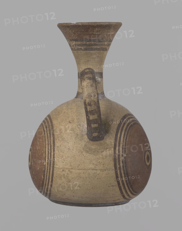 Cypriot jug, Jug on a low, straight foot. Globular body, flattened at the bottom. Quite narrow neck, funnel-shaped spout. Slightly flattened handle attached to the base of the neck and the upper part of the body. Painted decoration in stripes and linear patterns, in red-brown and black fading to brown. Near the spout is a wide red band, below it three black lines, the first adjacent to the red band. At the lower part of the neck, above the handle attachment, a narrow black band; another narrow black band at the base of the neck. The belly is decorated with three identical shooting target motifs on the front and sides: a wide red circular band with a black dot and a circle in the center, surrounded by four black-brown circles, the first adjacent to the red band. The handle is unpainted on the vessel side, black on the top, with light horizontal lines. The foot on the side is black. On the bottom, two concentric circles with a center marked with a dot. Good condition. Cypriot, Geometric ...