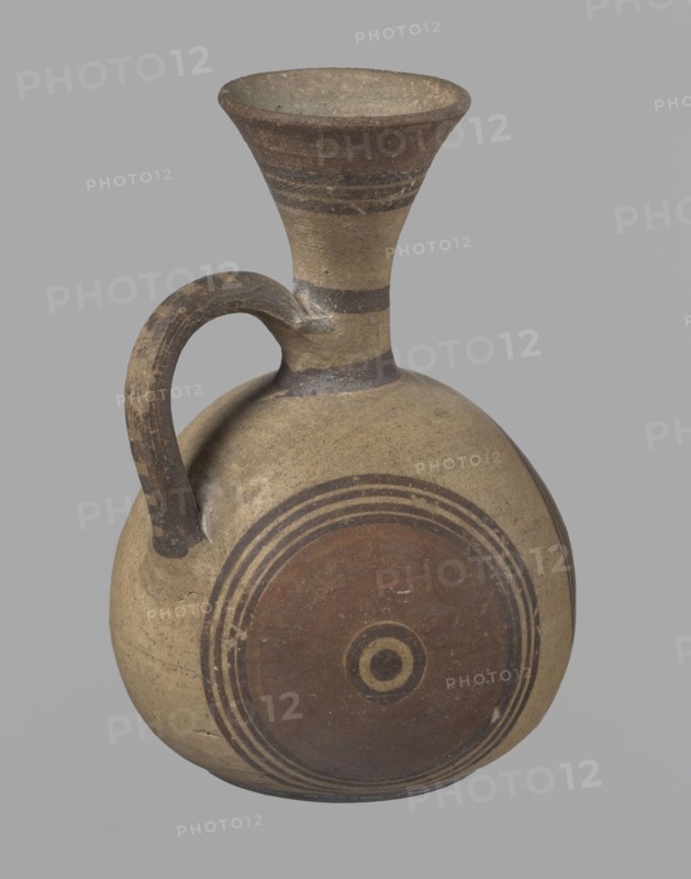 Cypriot jug, Jug on a low, straight foot. Globular body, flattened at the bottom. Quite narrow neck, funnel-shaped spout. Slightly flattened handle attached to the base of the neck and the upper part of the body. Painted decoration in stripes and linear patterns, in red-brown and black fading to brown. Near the spout is a wide red band, below it three black lines, the first adjacent to the red band. At the lower part of the neck, above the handle attachment, a narrow black band; another narrow black band at the base of the neck. The belly is decorated with three identical shooting target motifs on the front and sides: a wide red circular band with a black dot and a circle in the center, surrounded on the outside by four black-brown circles, the first adjacent to the red band. The handle is unpainted on the vessel side, black on top, with light horizontal lines. The foot on the side is black. On the bottom, two concentric circles with a center marked with a dot. Good condition. Cypriot ...