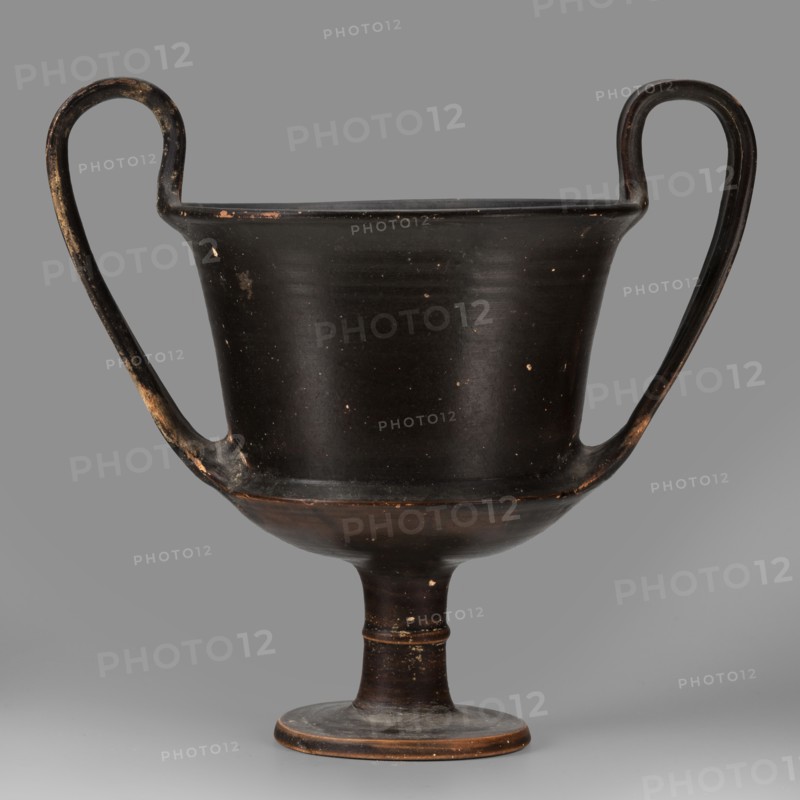 Kantharos, unknown Boeotian workshop. Physical description: Thin-walled kantharos with straight, slightly flared rim edges, a deep cylindrical bowl with an outer edge. The bowl is set on a high leg with a torus in the middle, flat foot. Band-like, wide handles with slightly raised edges, protruding above the level of the rim. The outer surface of the foot is slightly concave. Decoration: Vessel entirely covered with black varnish. Technology: Matte varnish, good quality, evenly distributed. Traces of turning visible under the varnish. Notes: Numerous secondary losses visible on the surface. Attributed to Kamila Nocon; 2nd half of the 5th century BC, Boeotia. Type: kantharos. Weight: 0.552 kg., A dark vessel with a footed base and a wide bowl rising to a flared rim, featuring a pair of high curved handles that extend from the rim down to the lower bowl; the surface shows small worn spots and areas of lighter wear. The predominant color is a deep blackish-brown with patches of lighter br...