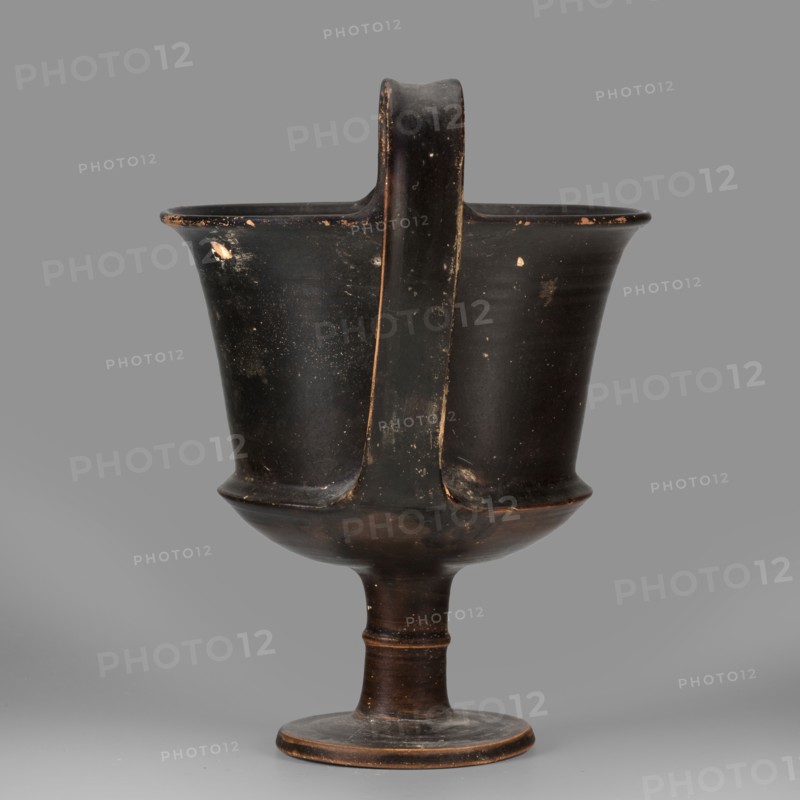 Kantharos, unknown Boeotian workshop. Physical description: Thin-walled kantharos with straight, slightly flared rim edges, a deep cylindrical bowl with an outer edge. Bowl set on a high leg with a torus in the middle, flat foot. Band-like, wide handles vices with slightly raised edges, protruding above the level of the rim. Outer surface of the foot slightly concave. Decoration: vessel entirely covered with black varnish. Technology: matte varnish, good quality, evenly distributed; traces of turning visible under the varnish. Notes: numerous secondary losses visible on the surface. Attributed to Boeotia, 2nd half of the 5th century BC. Weight 0.552 kg. Kamila Nocon, A dark, matte ceramic vessel with a tall, rounded bowl on a narrow stem and circular foot, featuring a single broad handle running from rim to base; the surface shows worn and chipped areas revealing lighter underlying tones, with small flecks and scratches; visible colors include black, dark brown, lighter brown from worn...