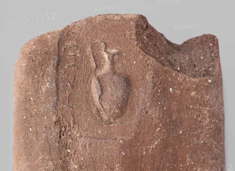 Fragment of a transport amphora vice with a stamp depicting an oinochoe and an incomplete Greek inscription. Fragment of an amphora vice with a rectangular stamp containing an inscription in two rows and between them a depiction of an oinochoe in a horizontal position. End of the 4th–beginning of the 3rd century BC. Found at Myrmekion; origin Thasos. Stamp; vice. Height 5.1 cm, width 10.8 cm., A weathered stone fragment in shades of brown and reddish-brown with a rough, pitted surface and small pale flecks scattered across it, featuring a shallow carved relief of an amphora-like vessel with a rounded body and two handles, a curved ridge along the top edge, and worn irregular edges that show chips and breaks.