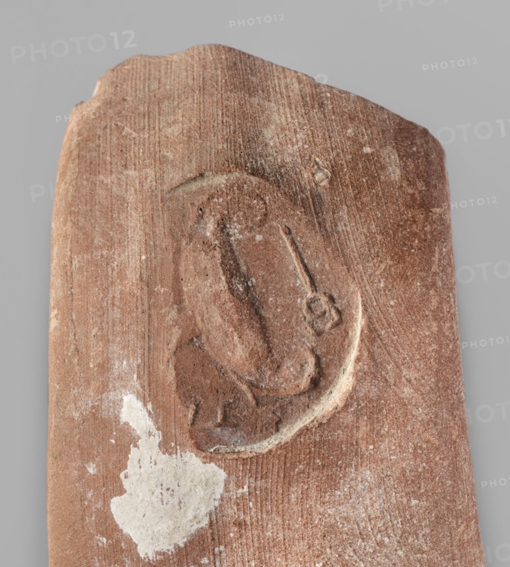 Fragment of an amphora with a stamp, ca. 390 BC, found at Pantikapaion, originating from Thasos; stamp; vice; height 6.2 cm, width 8 cm., A worn rectangular fragment with an uneven top edge in shades of reddish-brown with lighter brown and tan variations, showing a shallow oval impression carved into its surface that contains curved and rounded raised areas and a narrow linear indentation ending in a small circular cluster; there is a patch of rough white residue near the lower left of the fragment and faint lighter streaks and speckles across the surface, all set against a flat neutral gray background.