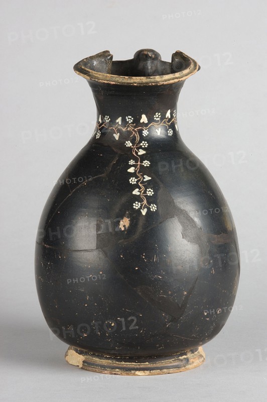 Oinochoe, Gnathia style; decoration: on the neck an engraved horizontal ivy sprig with white-painted heart-shaped leaves and flowers in the shape of pointed rosettes, and a second one extending downwards from it, placed centrally at the front; 2nd half of the 4th century BC, Apulia; oinochoe vessel; height 19.5 cm, width 13 cm., A rounded ceramic vessel with a narrow neck and flared rim, predominantly black in color with areas of worn, exposed lighter clay; a delicate vertical vine-like decoration in a pale color runs down from the neck with small heart-shaped and cluster-shaped leaf motifs; the rim shows chipped edges with a beige exposed layer and the base has a worn bead-like foot with patches of beige and darker paint; the surface of the vessel has a slightly glossy finish with scattered small chips and scratches revealing lighter tones beneath, and the background is a neutral light gray.