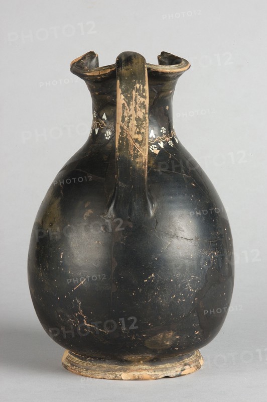Oinochoe, Gnathia style; decoration: on the neck an engraved horizontal ivy sprig with white-painted heart-shaped leaves and flowers shaped as pointed rosettes, and a second sprig extending downward from it placed centrally at the front; 2nd half of the 4th century BC; Apulia; oinochoe vessel; height 19.5 cm, width 13 cm., A single rounded ceramic vessel with a narrow neck and flared, slightly scalloped rim is shown against a plain light gray background, the surface of the vessel predominantly black with areas of worn beige where the finish has chipped away; a central handle runs from the rim to the shoulder and shows more extensive beige wear, and a small decorative band of lighter colored floral-like shapes sits near the neck; the base is slightly raised and shows exposed beige ceramic and wear, while the body displays a few scuffs and scratches revealing underlying lighter tones.