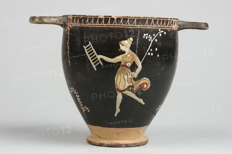 Skyphos, Skyphos with painted decoration. Engraved ornament at the upper edge of the vessel: an egg-shaped structure and a wavy line, framed by parallel lines. At the handles and in the center, vertical, engraved branches with preserved traces of white leaves hang downwards. Side A: a woman shown in left profile with a xylophone in her right hand and a branch in her left. The figure is flanked by small trees on either side. 4th century BC, Apulia, Southern Italy. Skyphos vessel, height 12.1 cm, width 11.1 cm., A black ceramic vessel with a rounded body and a narrow tan base, featuring two short horizontal handles at the rim; painted on the body is a pale-skinned figure in a short skirt and a darker sash, shown in a dynamic pose with one arm extended holding a ladder-like object and the other arm bent while holding a long slender stem topped with small pale dots, with pale decorative motifs around the figure and a patterned border near the rim in tan and brown tones.