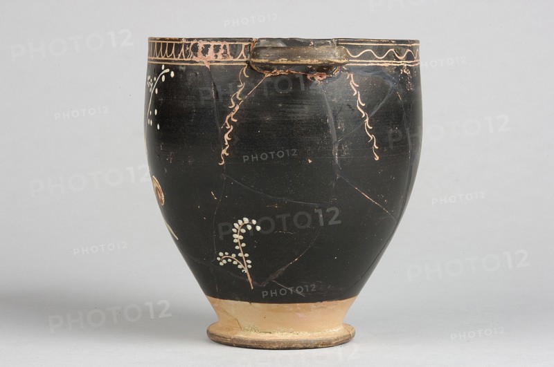 Skyphos with painted decoration. Engraved ornament at the upper edge: an egg-shaped motif and a wavy line, framed by parallel lines. At the handles and in the center, vertical engraved branches with preserved traces of white leaves hang downwards. Side A: a woman shown in left profile with a xylophone in her right hand and a branch in her left. The figure is flanked by small trees on either side. 4th century BC, Apulia, Southern Italy. Skyphos vessel, height 12.1 cm, width 11.1 cm., A dark rounded ceramic vessel with a small rim and a narrow base sits against a pale background, showing a predominantly black surface with areas of worn lighter brown and tan, and decorative pale peach squiggles and small pale dots forming simple botanical motifs; the base and foot are tan and light brown with some greenish residue, and there are visible fine cracks and chipped edges on the rim and body.