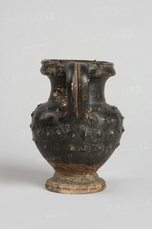 Black-finned oinochoe, Decoration: in the upper half of the belly 4 rows of plastic knobs applied using the barbotine technique. IV waC, Campania, vessel; height 9.8 cm, width 6.7 cm, The image shows a dark-colored ceramic jug with a rounded body, a narrow neck, a flared rim, and a single thick handle attached from the rim to the shoulder; the body has a textured surface with raised circular bumps and areas where the dark surface is worn away revealing lighter underlying clay, and the base shows a worn, earthy color. The visible colors include black, beige, brown, and hints of gray.