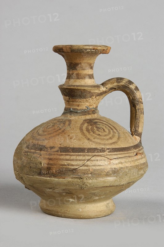 Jug, painted decoration: in the widest part of the body, on the neck and handle - horizontal lines of various widths; on the shoulders - 4 groups of 4 concentric circles; on the spout - concentric lines and small semicircles; 850–750 BC; Cypriot, Geometric Period III, Cyprus; vessel; height 7.5 cm, width 6.8 cm, A single ceramic jug with a rounded body, narrow neck, flared rim, and a curved handle is shown against a plain light gray background. The jug displays concentric circular patterns and horizontal bands in darker earth tones on a base of light beige clay; the colors visible include beige, tan, brown, and hints of ochre. The surface shows areas of wear and small chips, with a visible crack and some rough, textured patches where the clay appears abraded or repaired. The handle and neck have alternating lighter and darker horizontal stripes, and the rim has worn darker edges. The jug rests on a short footed base that is slightly lighter in color than the body.