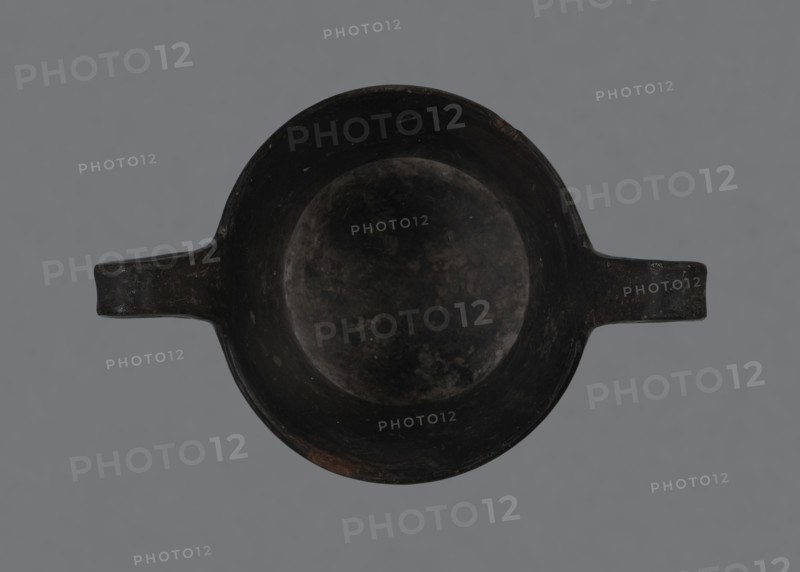 Kantharos, kantharos black-varnished on a cup-shaped foot, vessel; 1st half of the 6th century BC, Etruria Italy; function: goblet for banquets; provenance notes: repossession Bytom., A top-down view of a round, shallow bowl with a broad rim and a pair of short horizontal handles extending opposite each other, all appearing dark and worn with mottled surface textures; the background is a flat neutral gray. The visible colors are black, dark gray, and a few subtle brownish patches.
