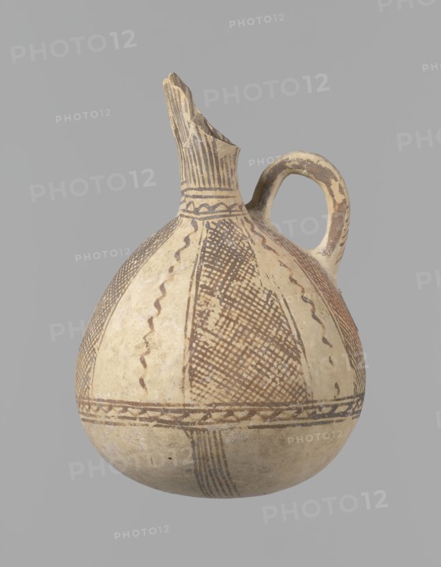 Jug, unknown Cypriot workshop. White painted ware Bronze Age jug with a spherical bottom and a bulging, pear-shaped body transitioning in the upper part into two tubular spouts with protruding walls. A ring-shaped vise protrudes between the spouts. Covered with white paint and decorated with brownish-red paint: a zigzag ornament runs around the body for one-third of its height, flanked on each side by two horizontal lines; below are four bands of lines converging at the bottom. The upper body has four stripes filled with diagonal, intersecting lines, separated by serpentine winding lines. The vise is decorated with diagonal lines at the top and brown paint on the sides. At the base of the rims, a zigzag ornament is framed by two lines on either side; vertical lines surround the rims both inside and outside. Date: 1900–1650 BC. Origin: Cyprus. Vessel; height 15 cm, width 13 cm., An off-white rounded ceramic vessel with a narrow neck and broken spout, featuring a single curved handle; th...