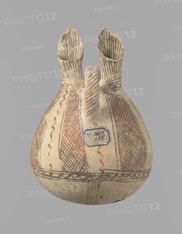 Jug, unknown Cypriot workshop. White painted ware jug with a spherical bottom, a bulging pear-shaped body transitioning in the upper part into two tubular spouts with protruding walls. Between them, a ring-shaped vise protrudes from the body. The vessel is covered with white paint and decorated with brownish-red paint. A zigzag ornament runs around the body for 1/3 of the vessel's height, flanked on each side by two horizontal lines. Below are four bands of lines converging at the bottom. The upper part of the body is decorated with four stripes filled with diagonal, intersecting lines. The stripes are separated by lines that wind serpentine. The vise is decorated with diagonal lines at the top and brown paint on the sides. At the base of the rims, a zigzag ornament is framed by two lines on either side; above it, vertical lines surround the rims both inside and outside. Bronze Age, 1900–1650 BC, Cyprus, vessel. Height 15 cm, width 13 cm., A rounded ceramic vessel with a bulbous body a...