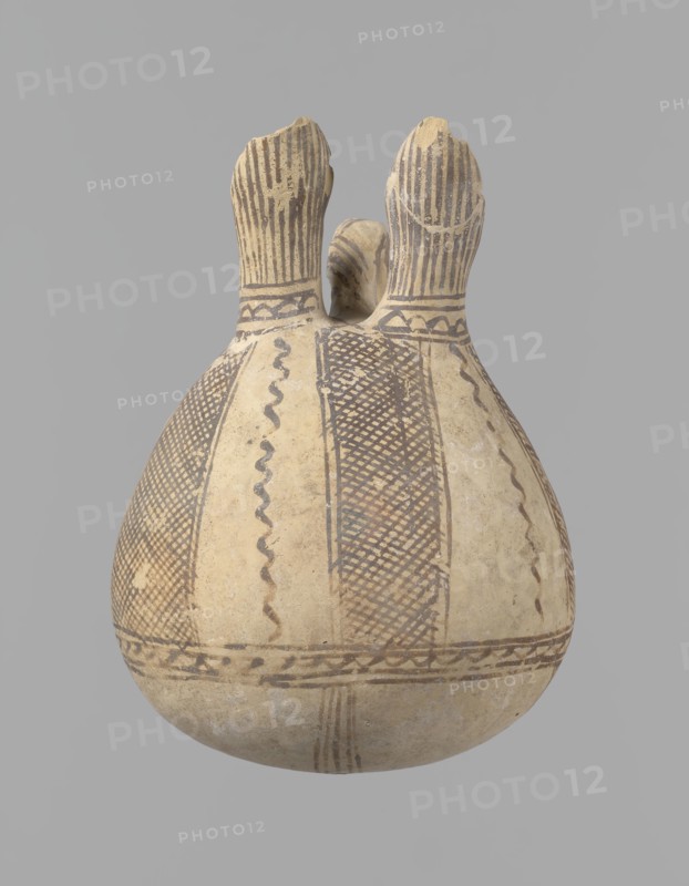 Jug, unknown Cypriot workshop, Cyprus, 1900–1650 BC. Vessel White painted ware with spherical bottom and bulging pear-shaped body transitioning in the upper part into two tubular spouts with protruding walls; a ring-shaped vise protrudes between them. Covered with white paint and decorated with brownish-red paint. A zigzag ornament runs around the body for one-third of the vessel's height, flanked on each side by two horizontal lines; below are four bands of lines converging at the bottom. The upper part of the body has four stripes filled with diagonal, intersecting lines, separated by serpentine winding lines. The vise is decorated with diagonal lines at the top and brown paint on the sides. At the base of the rims, a zigzag ornament is framed by two lines on either side; above it, vertical lines surround the rims inside and outside. Bronze Age jug, height 15 cm, width 13 cm., An off-white rounded ceramic vessel with a bulbous body and two upward-extending necks, decorated with hand-...