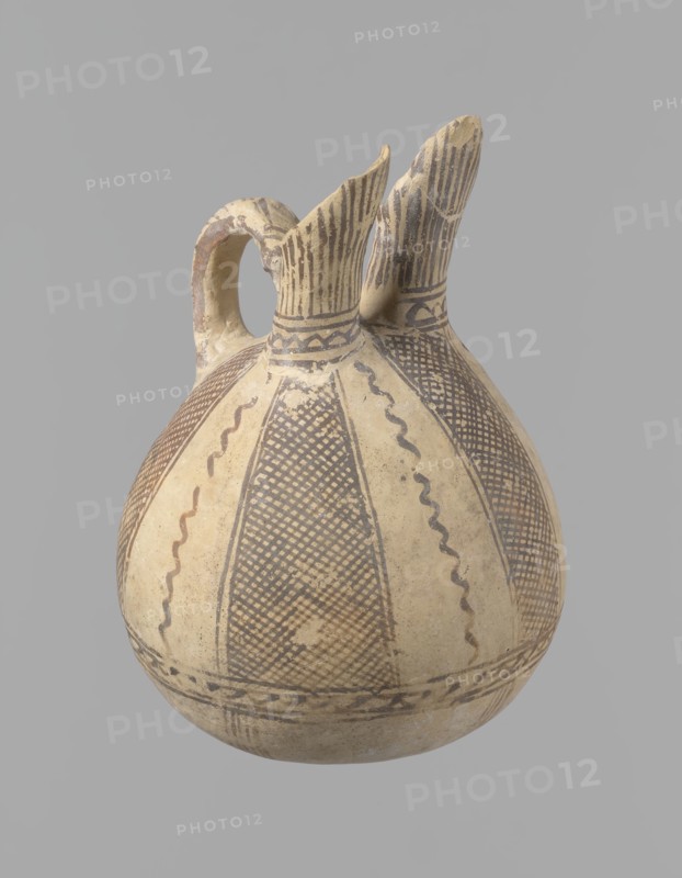 Jug, unknown Cypriot workshop. White painted ware jug with a spherical bottom, a bulging pear-shaped body transitioning in the upper part into two tubular spouts with protruding walls. Between them, a ring-shaped vise protrudes from the body. The vessel is covered with white paint and decorated with brownish-red paint. A zigzag ornament runs around the body for 1/3 of the vessel's height, flanked on each side by two horizontal lines. Below are four bands of lines converging at the bottom. The upper part of the body is decorated with four stripes filled with diagonal, intersecting lines; the stripes are separated by lines that wind serpentine. The vise is decorated with diagonal lines at the top and brown paint on the sides. At the base of the rims, a zigzag ornament is framed by two lines on either side; above it, vertical lines surround the rims, both inside and outside. Bronze Age, 1900–1650 BC, Cyprus. Vessel; height 15 cm, width 13 cm., A rounded ceramic vessel with a curved handle...