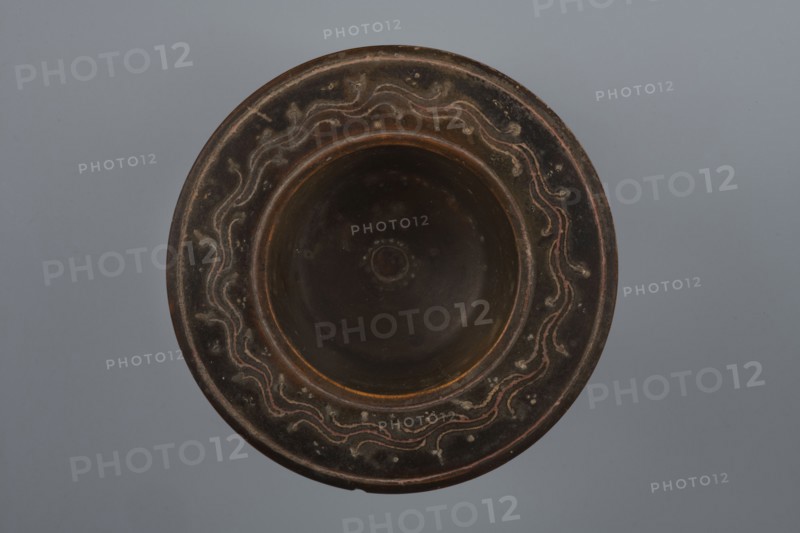 Black-finished bowl, 4th century BC, Southern Italy; vessel bowl; height 6.0 cm, width 11.5 cm., A round shallow bowl viewed from above with a wide rim and a recessed center, showing a dark brown to black surface with lighter brown and reddish accents; the rim features a repeating wavy decorative pattern in lighter beige and reddish tones against the darker background, and there are small raised dots and worn spots around the rim; the central recessed area is darker and smoother with subtle circular marks, and the background behind the object is a uniform soft gray.