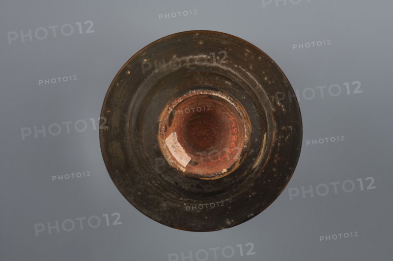 Black-finished bowl, 4th century BC, Southern Italy; vessel bowl; height 6.0 cm, width 11.5 cm., A top-down view of a circular object resembling a bowl or vessel with a wide rim and a recessed center, showing a worn, textured surface in dark brown and black tones with patches of lighter brown and ochre; the central interior has reddish-brown and terracotta hues with a few areas of chipped lighter beige and pale tan; the background is a smooth gray-blue.