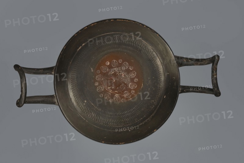 Black-finished goblet, 4th century BC, Campania; vessel goblet; height 5.0 cm, width 12.5 cm., A top-down view of a shallow round dish with two opposing handles, showing a predominantly dark surface with a central circular area in a reddish-brown hue. The rim and handles are dark, almost black, with small worn spots revealing lighter tones. Around the central reddish-brown area is a pattern of raised petal-like shapes and concentric textured bands in dark and slightly lighter shades. The background behind the dish is a muted gray.