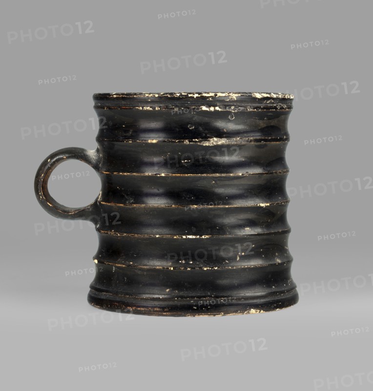 Dice cup. Small cup with a flat slightly concave bottom, slightly tapered walls, distinct rim and foot. Outer surface decorated with seven wide ribs; bottom has concentric decoration. Entire object covered with black varnish shiny but matte in places; traces of turning visible under varnish. Spout slightly chipped; surface shows dirt and secondary losses; varnish worn on rib ridges. Clay color 5 YR 6/6 reddish yellow; varnish color 2.5 YR 2.5/1 black. Dice game. 5th century BC, Attica. Vessel cup. Height 8.3 cm; width 10.6 cm; depth 8.3 cm., A worn black ceramic mug with a round handle on one side, featuring a ribbed, undulating surface with a few chips and worn areas revealing lighter material beneath; the background is a flat gray.