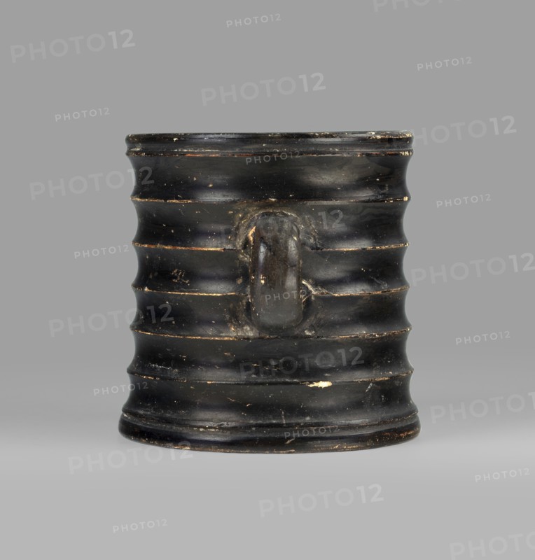 Dice cup. Small cup with flat slightly concave bottom; slightly tapered walls; distinct rim and distinct foot. Outer surface decorated with seven wide ribs. Entire object covered with black varnish shiny but in places matte, except for a small fragment on the inner surface and the outer surface of the bottom, which has a concentric decoration. Traces of turning visible under the varnish. Spout slightly chipped; surface shows dirt and secondary losses; varnish worn on rib ridges. Clay color: 5 YR 6/6 reddish yellow. Varnish color: 2.5 YR 2.5/1 black. 5th century BC, Attica. Vessel cup. Height 8.3 cm; width 10.6 cm; depth 8.3 cm., A ribbed cylindrical vessel with a single rounded handle projecting from its side, shown against a neutral gray background; the vessel is predominantly black with small areas of worn lighter brown and off-white showing through its surface, and it has a slightly glossy finish with visible scuffs and chips.