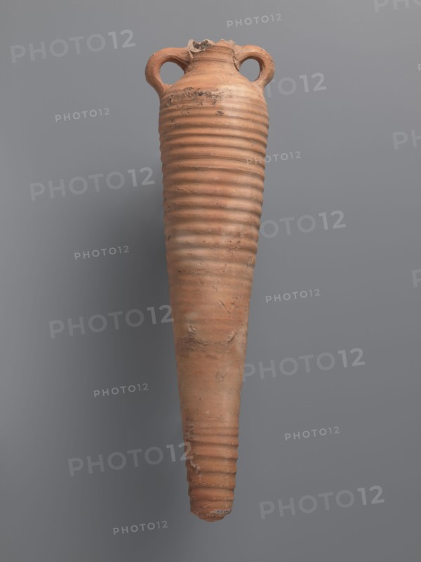 Amphora, unknown Aswan workshop. Spindle-shaped amphora made of pink kaolin clay Gempeler clay IA, 6th–7th century. Thick, ring-shaped vises protruding beyond the vessel outline are attached to the base of the neck and the lower part of the arms. The upper half and lowest section have a fluted surface traces of turning. The bottom is conical, flat-topped. Outer surface smoothed and covered with red engobe, in places burnt to a cream-orange color. Interior coated with black resin impregnation. Material: kaolin clay. Function: storage vessel Coptic amphora. Find location: Edfu. Dimensions: height 50.8+ cm preserved; neck diameter 5.5 cm; body diameter 12.4 cm., A tall, elongated ceramic vessel with a narrow pointed base and a wider rounded top, featuring a ribbed texture along most of its body and a rough, chipped rim with two looped handles near the top; the surface shows patches of wear and uneven texture. The visible colors are terracotta, brown, beige, gray, and muted orange, set aga...