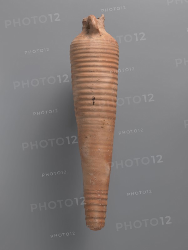 Amphora, unknown Aswan workshop. Spindle-shaped amphora made of pink kaolin clay Gempeler clay IA, 6th–7th century. Thick, ring-shaped vises protrude beyond the outline of the vessel and are attached to the base of the neck and the lower part of the arms. Upper half and lowest section have a fluted surface traces of turning. Conical, flat-topped bottom. Outer surface smoothed and covered with red engobe, in places burnt to a cream-orange color. Interior coated with black resin impregnation. Find location: Edfu. Vessel amphora, height 50.8+ cm preserved, neck diameter 5.5 cm, body diameter 12.4 cm., A tall, tapered clay vessel with a pointed bottom and a narrower top, featuring horizontal ribbed bands running along its length; the surface shows rough texture and small imperfections, with a broken or irregular rim at the top. The visible colors are terracotta, light brown, and hints of darker brown against a neutral gray background.