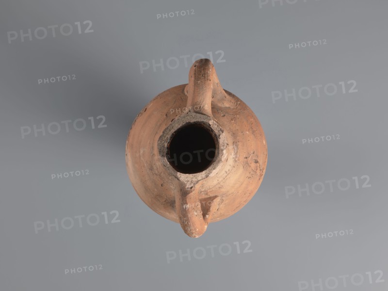 Spindle-shaped amphora, 6th–7th century. Made of pink kaolin clay Gempeler clay IA. Thick, ring-shaped vises protruding beyond the outline are attached to the base of the neck and the lower part of the handles. Upper half and lowest section have a fluted surface traces of turning. Conical, flat-topped bottom. Outer surface smoothed and covered with red engobe, in places burned to a cream-orange color. Interior coated with black resin impregnation. Amphorae type associated with an Aswan workshop; Egyptian/Coptic ceramic storage vessel. Find location: Edfu. Preserved height 50.8+ cm; neck diameter 5.5 cm; body diameter 12.4 cm., A top-down view of a round clay vessel with a central wide opening and a handle-like projection, showing a weathered surface with visible rough textures and chips; the vessel is predominantly terracotta with shades of brown and orange, and the background is a smooth gray.