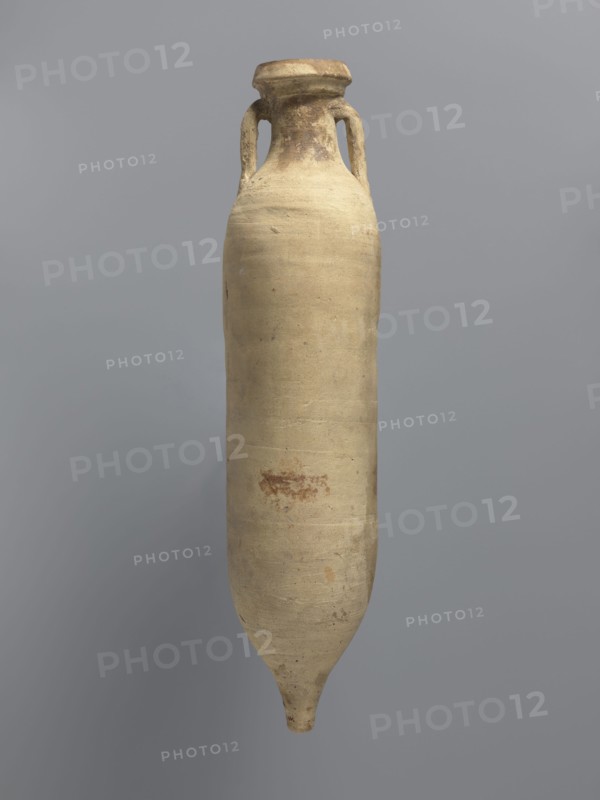 Amphora spatheion, unknown North African workshop Tunisia/Libya. Cylindrical, elongated amphora with a smooth yellow-green surface, probably covered with a coating produced during firing natural engobe, so-called self-slip. Two-stage profiled spout with a rounded top and an outward-sloping lip. Small loop-shaped handles glued to the arms and to the neck just below the spout; conical bottom. Inner walls covered with a resin impregnation, also visible on the outer walls of the upper part of the amphora. Complete vessel, ceramic body not visible. 2nd half of the 6th–7th century; findspot: Edfu. Vessel amphora. Height 44.2 cm; diameter 6.6 cm rim, 10 cm body., A tall, elongated ceramic vessel with a narrow neck and a small rounded foot, showing a pair of looped handles near the top; the surface is mostly beige with areas of darker brown and light tan, and there are a few small reddish-brown spots and subtle variations in texture on the body, set against a smooth gray background.